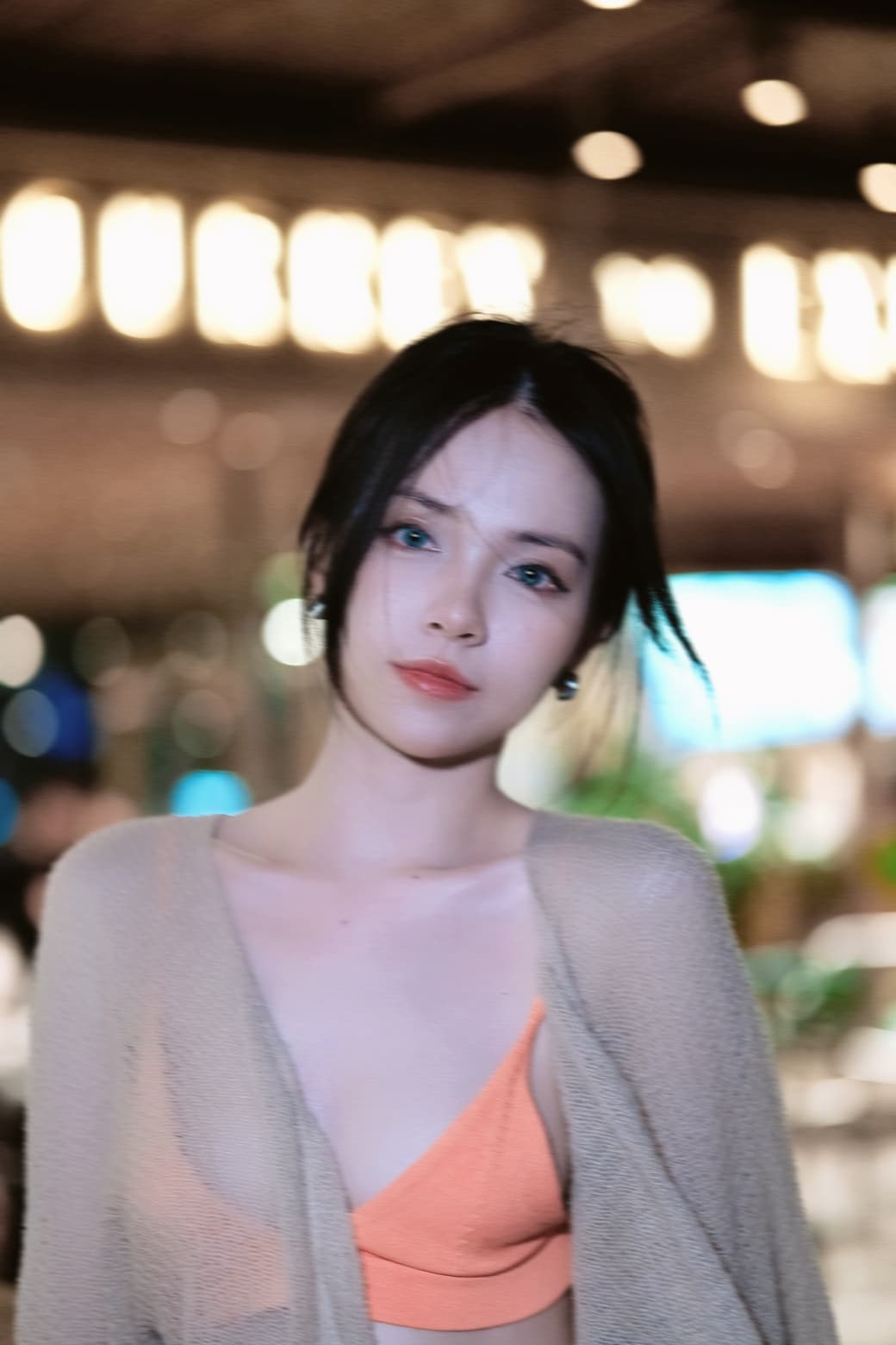 A woman with dark hair, wearing a light-colored shawl over an orange top, stands in a softly lit indoor setting with blurred lights in the background