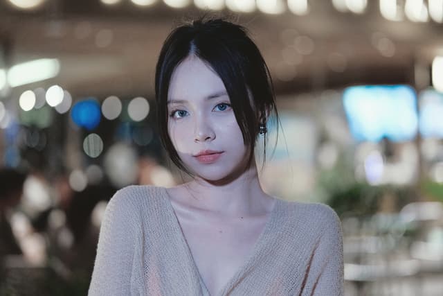 A woman with long dark hair, wearing a light-colored top, standing in a softly lit indoor setting with blurred lights and people in the background