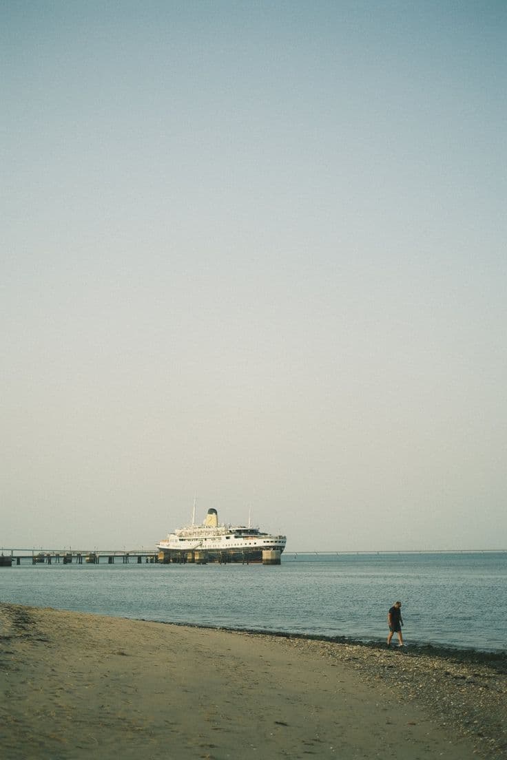 A large ship docked at a pier with a person standing on a sandy beach in the foreground, under a clear sky